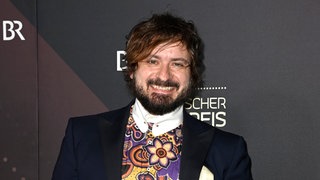 Schauspieler Daniel Christensen steht vor einer Wand und trägt einen Anzug. Er lacht direkt in die Kamera