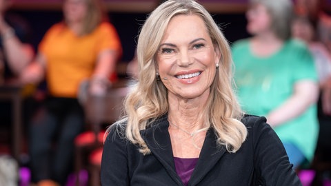 Nina Ruge sitzt auf einem Stuhl mit einem schwarzen Sakko und lacht in die Kamera.