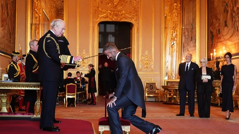 David Beckham wird von König Charles in einem festlichen Saal auf Schloss Windsor zum Ritter geschlag. Seine Eltern und Partnerin Victoria Beckham sind im Hintergrund zu sehen.