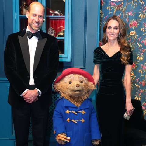 Prinzessin Kate und Prinz William haben elegante Kleider an bei der Royal Variety Performance 2025. Das roylare Paar steht neben dem Bär Paddington und lächelt.
