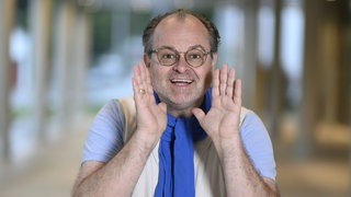 Schauspieler Markus Majowski trägt einen blauen Schal und lacht direkt in die Kamera und hat beide Hände neben dem Gesicht.