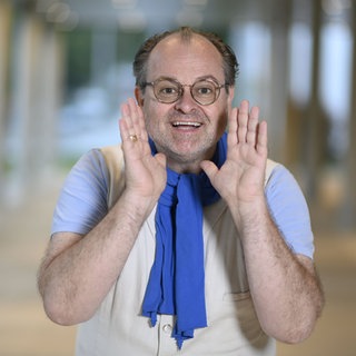 Schauspieler Markus Majowski trägt einen blauen Schal und lacht direkt in die Kamera und hat beide Hände neben dem Gesicht.