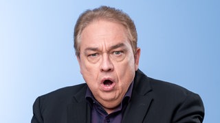 Oliver Kalkofe im schwarzen Sakko hebt die Hände und schaut erschrocken in die Kamera.