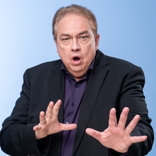 Oliver Kalkofe im schwarzen Sakko hebt die Hände und schaut erschrocken in die Kamera.