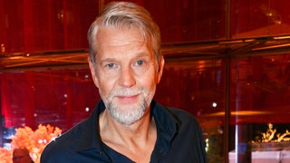 Schauspieler Kai Wiesinger steht in Hemd und Jeans in einer Lobby auf einem roten Teppich und schaut neutral bis leicht ernst in die Kamera.