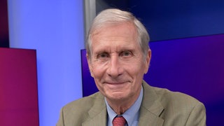 Ulrich Wickert trägt einen beigen Anzug und eine rote Krawatte und schaut direkt in die Kamera und lächelt.