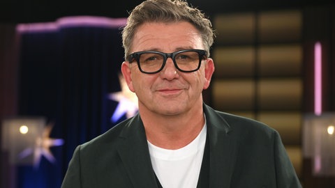 Schauspieler Hans Sigl trägt eine schwarze Brille und schaut direkt in die Kamera.