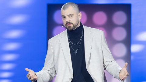 Comedian Nikita Miller trägt bei einem Auftritt ein hellgraues Sakko und ein schwarzes Unterteil mit einer silbernen Kette.