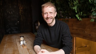 Ein junger Mann, David Zimmerschied, mit rötlichen kurzen Haaren sitzt in schwarzem Pulli an einem Holztisch und lächelt.