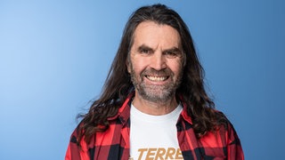 Der Extrembergsteiger Thomas Huber steht vor einem blauen Hintergrund und lächelt in die Kamera.