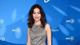 Sabrina Amali steht im Kostüm vor einer ARD Pressewand und lächelt