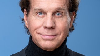 "Der Alte"-Schauspieler Thomas Heinze vor blauem Hintergrund mit schwarzem Jacket. Er schaut direkt in die Kamera.