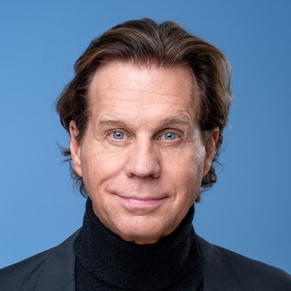 "Der Alte"-Schauspieler Thomas Heinze vor blauem Hintergrund mit schwarzem Jacket. Er schaut direkt in die Kamera.