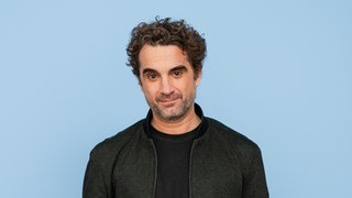Der Schauspieler Oliver Wnuk trägt ein schwarzes Shirt und eine schwarze Jacke vor blauem Hintergrund und schaut direkt in die Kamera.