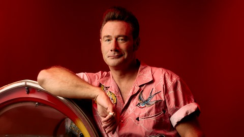 Dick Brave steht neben der Jukebox und lehnt sich mit dem Arm daran ab.