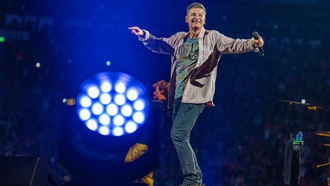 Hartmut Engler steht auf der Bühne, streckt die Arme aus und hat in einer Hand ein Mikrofon