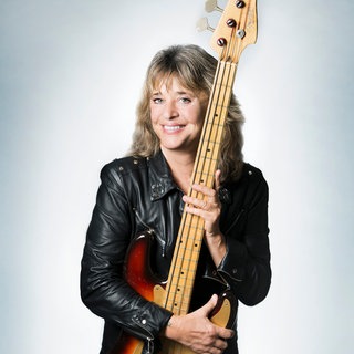 Sängerin Suzi Quatro steht in einer Lederjacke und einer Gitarre in der Hand und schaut direkt in die Kamera