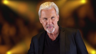 Johnny Logan steht in einem schwarzen Anzug gegen die Wand gelehnt und schaut direkt in die Kamera