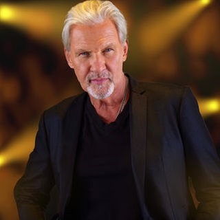 Johnny Logan steht in einem schwarzen Anzug gegen die Wand gelehnt und schaut direkt in die Kamera
