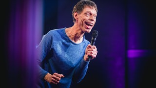 Comedian Maddin Schneider schaut von links ins Bild mit einem Mikro in der Hand und hat den Mund offen
