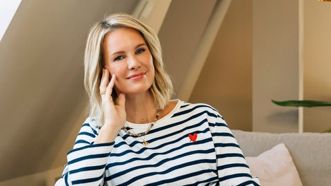 Monica Meier Ivancan sitzt mit angezogenen Beinen auf der Couch und trägt ein schwarz-weiß gestreiftes Oberteil und eine blaue Hose