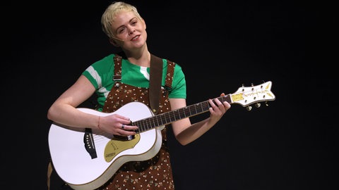 Jennifer Siemann, in der Rolle als Nina Bauer im Musical "Ein bisschen Frieden", steht mit einer weißen Gitarre auf der Bühne und singt. 