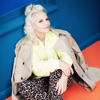 Ina Müller sitzt auf dem Boden in gelbem Oberteil und brauner Jacke über den Schultern