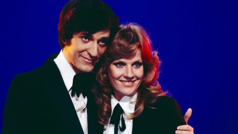 "disco" Moderator Ilja Richter posiert mit Sängerin und Schauspielerin Heidi Brühl vor einem blauen Hintergrund und hält sie im Arm. Bekannt wurde er auch durch seine Sketche und die "Pauker"-Filme.