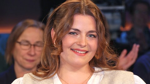 Ronja Forcher sitzt in einem Stuhl und hat eine weiße Bluse und eine beige Weste an