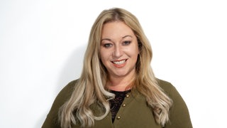 Rebecca Siemoneit-Barum steht vor einer weißen Wand mit grünen Strickjacke und Jeans und hat die Hände in den Hosentaschen