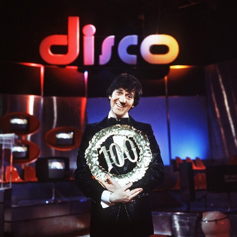 "disco"-Moderator Ilja Richter präsentiert die 100. Sendung des TV-Klassikers und steht vor einem dunklen Hintergrund mit einer bunten Leuchtschrift "disco". Er hält einen silbernen Kranz mit einer silbernen 100 in den Händen. Bekannt wurde er auch durch seine Sketche und die "Pauker"-Filme.