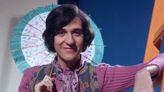 "disco" Moderator Ilja Richter lehnt an einer orangefarbenen Leiter trägt einen lilanen Pullover und schaut in die Kamera. Bekannt wurde er auch durch seine Sketche und die "Pauker"-Filme.