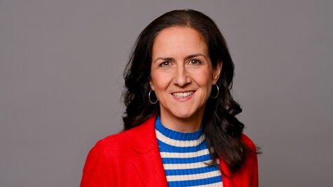 Die Moderatorin Chrissie Weiss lächelt in die Kamera. Sie trägt einen blauweiß-geringelten Pulli und darüber einen roten Blazer.