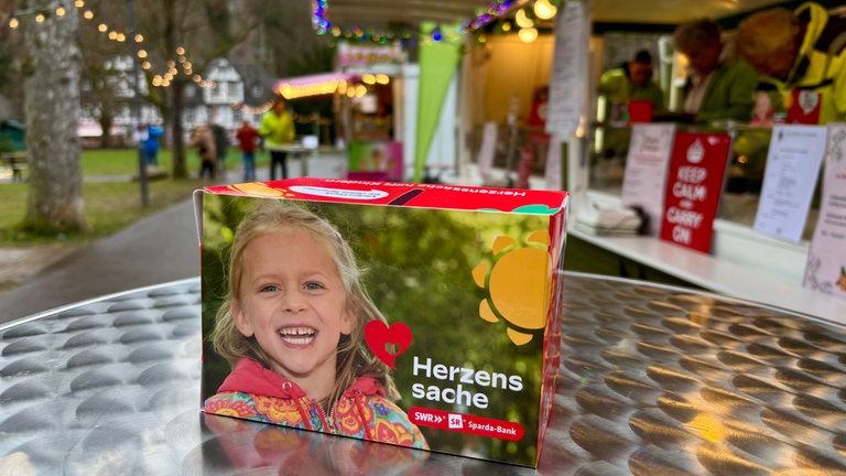 Auf einem Tisch neben dem grünen Adventsmobil von SWR4 steht eine rote Spendenbox der Herzenssache. 