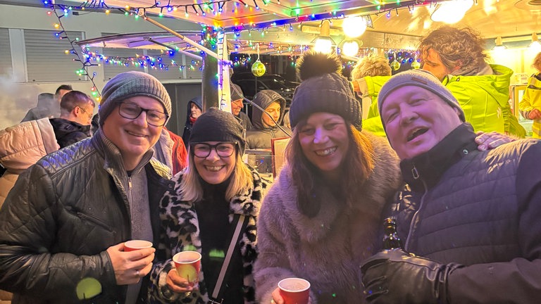 Weihnachtsmarkt in Ramstein: Eine lustige Gruppe trinkt Glühwein und lacht vor dem grünen SWR4 Adventmobil in die Kamera. 