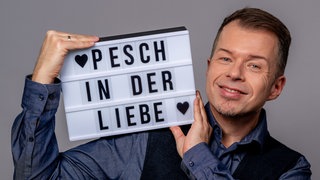 Moderator Daniel Pesch hält eine Lightbox mit dem Text "Pesch in der Liebe" hoch und lächelt.