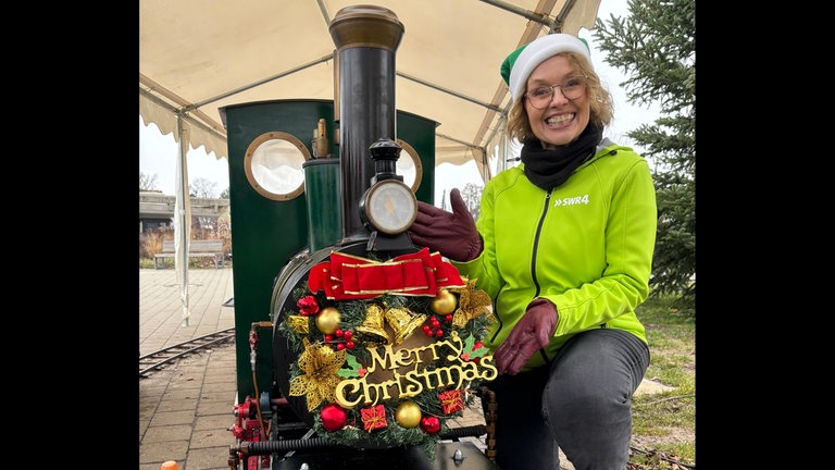 Weihnachtsmarkt in Fellbach: SWR4 Moderatorin Sabine Gronau kniet neben einer kleinen Dampflok, die weihnachtlich geschmückt ist.
