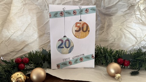 Eine verzierte, weiße Weihnachtskarte. In der Mitte sind zwei Christbaumkugeln, mit den Mustern eines 20 und 50 Euro Geldscheins, die an einem Dekoband hängen. Ein schöne Idee für eine Geldgeschenk an Weihnachten.