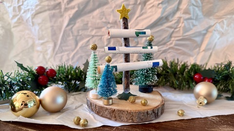 Auf eine Holzscheibe sind mehrer kleine Dekotannenbäume geklebt und in der Mitte ein längerer Stock, dessen Äste eingerollte Geldscheine sind. So schön können Geldgeschenke an Weihnachten sein.