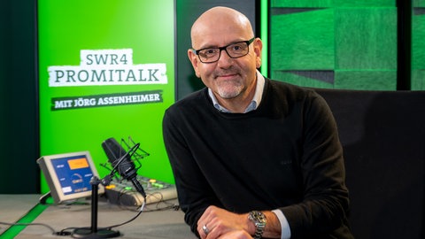 Moderator Jörg Assenheimer sitzt lächelnd im Studio am Tisch mit zwei Mikros und Bildschrim, im Hintergrund das Logo des SWR4 Promitalk auf einem Monitor.