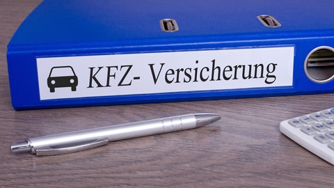 Ein blauer Aktenordner mit Aufschrift "KFZ-Versicherung" liegt auf dem Tisch.