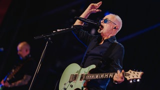 Nik Kershaw steht mit seiner Gitarre auf der Bühne und hält eine Hand nach oben