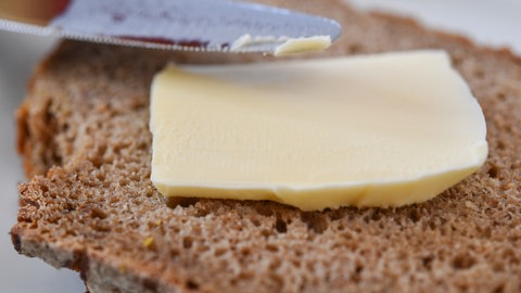 Ein Stück Butter liegt auf einer Scheibe Brot - ein Messer ist daneben zu sehen, um die Butter zu verstreichen.