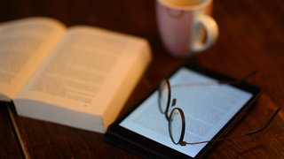 Ein Buch und ein iPad Tablet mit geöffneten eBook liegt auf einem Tisch (Symbolbild)
