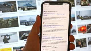 News auf einem Smartphone in einer Hand mit News zum Krieg in der Ukraine. Im Hintergrund Nachrichtenbilder, Bilder auf einem PC-Monitor zum Krieg in der Ukraine.