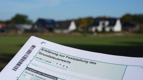Ein Erklärung zur Feststellung des Grundsteuerwerts für das Jahr 2025 wird vor eine Wohnsiedlung hochgehalten.