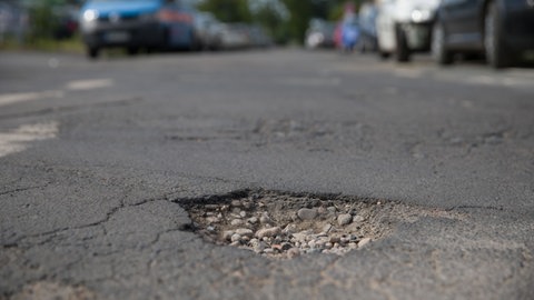 Mitten auf einer Straße ist ein Schlagloch zu sehen - im Hintergrund parken Autos am Straßenrand.