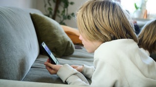 Ein Junge liegt auf einem Sofa und guckt auf sein Smartphone. (gestellte Szene)