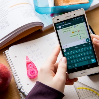 Eine Schülerin sitzt mit ihrem Handy und ihren Schulsachen an einem Schreibtisch und hat einen Chat geöffnet.