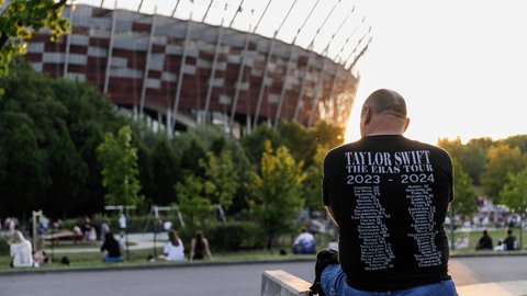 Ein Mann sitzt vor einem Stadion im Sonnenuntergang. Man sieht ihn von hinten, er trägt ein Shirt auf dem steht: Taylor Swift, THE ERAS TOUR 2023-2024